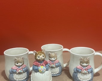 Vintage Otagiri "Mrs. Whiskers" ceramic cat bell and 3D relief mugs featuring kittens dressed in blue dresses, white aprons, and red bows.