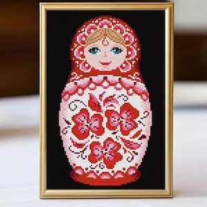 May include: Framed cross-stitch of a Matryoshka doll. The doll has a red headscarf and floral design on a white body. The doll has blue eyes and a smiling face. The frame is gold-coloured, and the background is black.