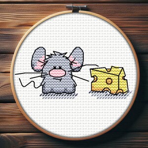 May include: Cross-stitch embroidery in a wooden hoop depicts a cartoon mouse with grey fur, pink ears, and a pink nose. The mouse is next to a yellow cheese wedge with holes. The background is white.