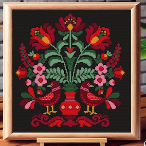 May include: A framed cross-stitch featuring a symmetrical floral design with two birds. The artwork uses red, pink, yellow, and green threads on a black background. The wooden frame adds a rustic touch to the piece.