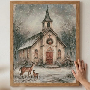 May include: A framed wall art print depicting a snowy winter scene with a church building featuring a steeple, a cross, and a wreath on the facade. Two deer stand in the foreground on snow-covered ground surrounded by bare trees and evergreens.