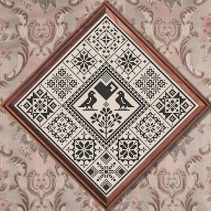May include: A square cross-stitch embroidery piece framed in a diamond orientation within a wooden frame. The design features black geometric patterns on a white background, including a central heart, two birds, and a floral motif surrounded by squares.