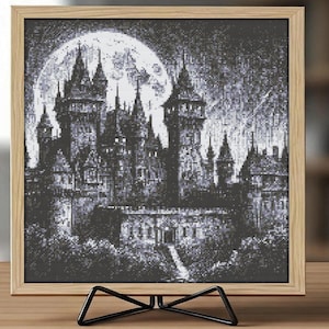 May include: A framed black and white cross-stitch of a gothic castle under a full moon. The artwork is displayed on a wooden stand. The castle features multiple spires and towers, with a detailed, pixelated design.