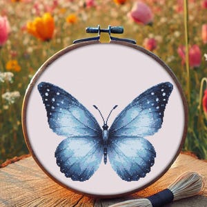 Op de afbeelding: Een blauwe vlinder met zwarte stippen op zijn vleugels is geborduurd op een wit canvas. Het canvas is gespannen op een houten borduurring.