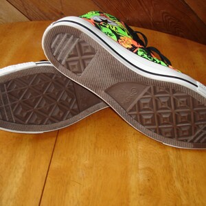 K&ouml;nnte beinhalten: Paar Converse All Star High-Top-Sneaker. Die Sneaker haben eine wei&szlig;e Gummisohle und ein schwarz-wei&szlig; gemustertes Obermaterial mit neongr&uuml;nen und orangefarbenen Akzenten. Die Sohlen sind braun mit einem Rautenmuster.