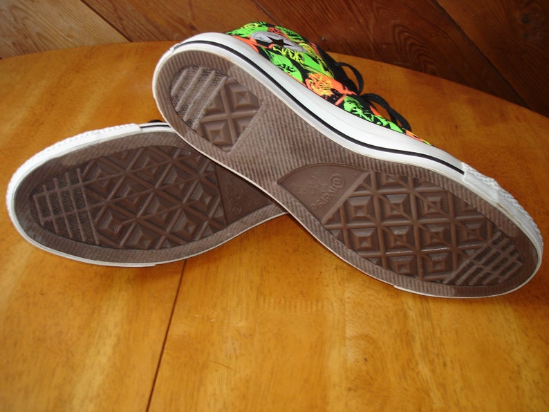 K&ouml;nnte beinhalten: Paar Converse All Star High-Top-Sneaker. Die Sneaker haben eine wei&szlig;e Gummisohle und ein schwarz-wei&szlig; gemustertes Obermaterial mit neongr&uuml;nen und orangefarbenen Akzenten. Die Sohlen sind braun mit einem Rautenmuster.