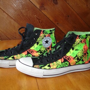 K&ouml;nnte beinhalten: Paar hohe Converse-Sneaker. Die schwarzen Canvas-Schuhe weisen ein lebendiges Muster aus neongr&uuml;nen und orangefarbenen Formen auf. Zunge und Schn&uuml;rsenkel sind schwarz, mit einer wei&szlig;en Zehenkappe und Sohle aus Gummi. Das Converse All Star-Logo befindet sich an der Seite.