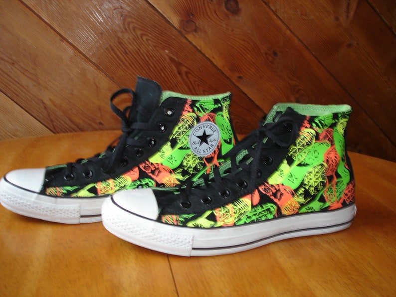 K&ouml;nnte beinhalten: Paar hohe Converse-Sneaker. Die schwarzen Canvas-Schuhe weisen ein lebendiges Muster aus neongr&uuml;nen und orangefarbenen Formen auf. Zunge und Schn&uuml;rsenkel sind schwarz, mit einer wei&szlig;en Zehenkappe und Sohle aus Gummi. Das Converse All Star-Logo befindet sich an der Seite.