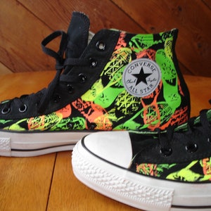K&ouml;nnte beinhalten: Schwarze High-Top-Sneaker mit einem leuchtenden neongr&uuml;nen und orangefarbenen Muster. Die Sneaker haben eine wei&szlig;e Gummikappe und Sohle, schwarze Schn&uuml;rsenkel und das Converse All Star-Logo.