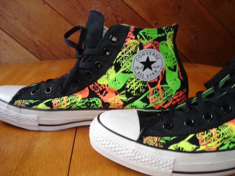 K&ouml;nnte beinhalten: Schwarze High-Top-Sneaker mit einem leuchtenden neongr&uuml;nen und orangefarbenen Muster. Die Sneaker haben eine wei&szlig;e Gummikappe und Sohle, schwarze Schn&uuml;rsenkel und das Converse All Star-Logo.