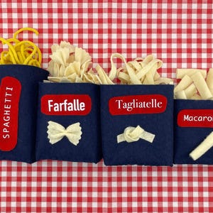 Könnte beinhalten: Vier marineblaue Filzbehälter gefüllt mit verschiedenen Nudelformen: Spaghetti, Farfalle, Tagliatelle und Makkaroni. Jeder Behälter hat ein rotes Etikett mit dem Pastanamen in Weiß. Der Hintergrund ist ein rot-weiß kariertes Muster.