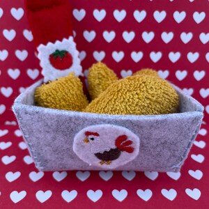 Op de afbeelding: Een grijze vilten mand met een haanapplicatie, gevuld met gele items en een rood vilten item met een tomatenontwerp. De achtergrond is rood met witte harten.