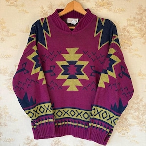 May include: A vintage magenta sweater with olive green and navy blue geometric patterns. The sweater has a V-neck and ribbed cuffs and hem. The design includes zigzag and diamond shapes. A retro knitwear piece.
