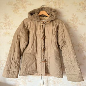 Peut inclure: Veste matelassée beige avec capuche doublée de fausse fourrure. Elle est dotée de fermetures à bascule en bois et de deux poches avant. La veste est suspendue à un cintre en bois sur un fond de papier peint à motifs.