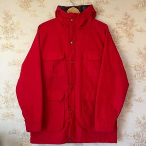 Puede incluir: Chaqueta roja brillante con capucha, cuatro bolsillos delanteros y cierre de botones. La chaqueta tiene un diseño clásico, adecuada para actividades al aire libre. La capucha está forrada con una tela estampada.