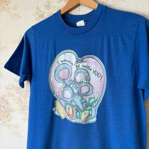 May include: A blue t-shirt with a graphic of a cartoon mouse inside a heart-shaped design. The text "I WANNA BE WITH YOU!" is printed around the mouse. The shirt is hanging on a wooden hanger.