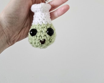 May include: A handmade crochet charm in the shape of a cartoon flask. The flask has a white top and a green body with large black eyes and a stitched smile. A black string is attached for hanging.