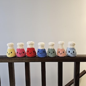 Puede incluir: Siete vasos de precipitados de ganchillo con cuerpos de diferentes colores y tapas blancas. Cada vaso de precipitados tiene una cara sonriente.