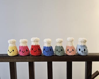 May include: Seven crocheted beakers with different colored bodies and white tops. Each beaker has a smiling face.