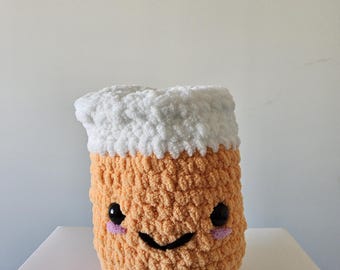 May include: A crocheted plush toy shaped like a beer mug with a white foam top. The mug has a smiling face with black eyes and pink cheeks.