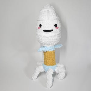 May include: A plush crochet bacteriophage toy featuring a large white head with black bead eyes and pink stitched cheeks, a light blue collar, a mustard yellow cylindrical midsection, and six white tentacle-like legs at the base.