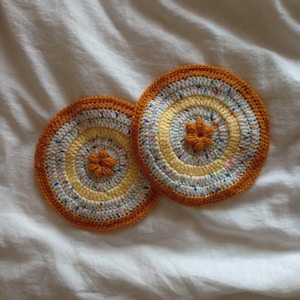 Puede incluir: Dos posavasos redondos hechos a mano con ganchillo. Cada posavasos presenta círculos concéntricos de hilo amarillo, blanco y moteado, con un borde naranja. En el centro de cada posavasos hay un pequeño adorno en forma de flor naranja.