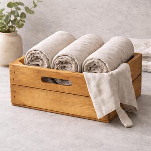 Op de afbeelding: Een houten kist met drie opgerolde beige linnen handdoeken. Een vierde handdoek is over de zijkant gedrapeerd. De kist is gemaakt van lichtbruin hout en heeft een handvat aan de voorkant. Een vaas met groen staat op de achtergrond.