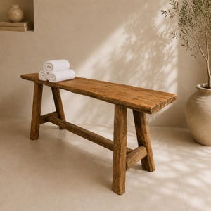 Op de afbeelding: Een rustieke houten bank met een natuurlijke afwerking. Drie opgerolde witte handdoeken liggen op de bank. De bank heeft een rechthoekige zitting en schuine poten. Het hout heeft zichtbare nerven en knopen.