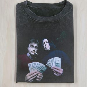 Może przedstawiać: Ciemnoszara koszulka z nadrukiem przedstawiającym dwóch mężczyzn trzymających pieniądze. Jeden mężczyzna nosi okulary i pali papierosa. Drugi mężczyzna jest w ciemnej szacie. Koszulka jest złożona.