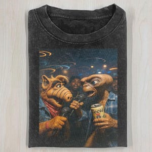 May include: Dark gray t-shirt featuring a humorous graphic of ALF and E.T. singing into microphones. ALF wears a denim jacket and a red bandana, while E.T. holds a Coors beer can. The image has a vintage, distressed look.