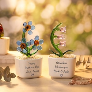 Könnte beinhalten: Zwei Glasblumenskulpturen in weißen quadratischen Töpfen. Eine zeigt blaue Blumen mit orangefarbenen Zentren, die andere rosa glockenförmige Blumen. Die Töpfe sind mit Botschaften und Herzen beschriftet, ideal für den Muttertag oder eine Großmutter.