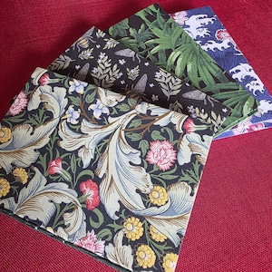 Può includere: Un set di tre quaderni rettangolari con copertine distinte. Il quaderno anteriore presenta uno sfondo scuro con foglie di acanto azzurre, fiori rosa e gialli. Dietro, una copertina nera mostra insetti e fogliame, mentre il quaderno posteriore presenta un motivo a foglie di palma verdi e un design a onde blu.