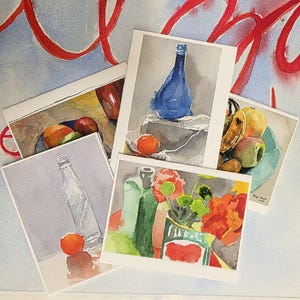May include: A collection of watercolor art prints featuring still life compositions. The prints showcase a blue bottle, fruit, and flowers. The color palette includes shades of blue, orange, green, and red. The artwork is displayed on a light background.
