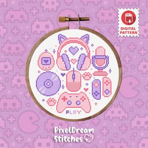 Puede incluir: Diseño de punto de cruz en un aro de madera, con iconos de videojuegos rosas y morados como auriculares, un ratón y un mando. El diseño incluye el texto "PixelDream Stitches" en la parte inferior. El fondo es un patrón repetitivo.