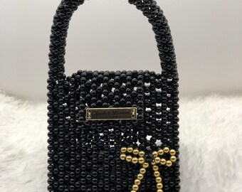 May include: A black beaded handbag with a rounded handle. The bag features a gold bow made of beads and a rectangular gold label that reads "Beads & Blossom". The bag is on a white, fluffy surface.