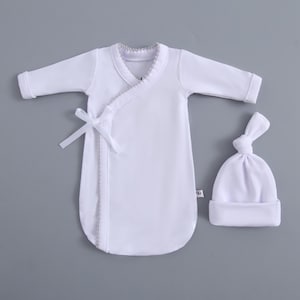 May include: A white baby sleep sack and matching knotted hat set. The sleep sack has long sleeves, a wrap-style front with a tie closure, and a decorative trim. The hat is a simple, soft design. The set is made of a soft material.