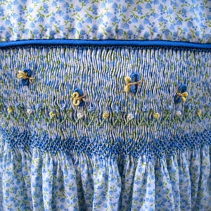 May include: Close-up of a blue and white floral patterned dress with smocked bodice. The bodice features embroidered butterflies in blue and yellow, with delicate green and white floral accents. A blue satin ribbon trims the neckline.