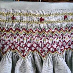 Può includere: Primo piano di una sezione di tessuto azzurro con ricami intricati. Il ricamo presenta file di filo dorato e rosso, creando un motivo strutturato. Piccoli accenti floreali rossi sono distribuiti uniformemente lungo la fila superiore, aggiungendo un tocco delicato al design.