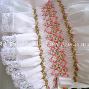 May include: Close-up of a white garment with intricate details. Features include smocking with coral and green embroidery, ruffled sleeves, and lace trim. A white satin ribbon with a small floral accent adds a delicate touch. The text "© EverRabbitDesigns.com" is visible.