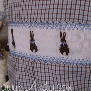 May include: A close-up of a white and blue checkered fabric with a decorative band featuring embroidered brown bunnies with blue bows. The design is reminiscent of Easter or spring themes.
