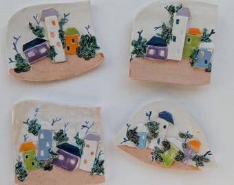 May include: Four ceramic wall hangings, each depicting a small village with houses in various colours, including purple, blue, green, and orange. The houses are surrounded by trees and have white roofs.