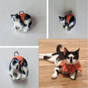 May include: A ceramic cat pendant with a black, white, and orange calico pattern. The pendant has a small loop at the top for attaching to a necklace or keychain. The cat is in a sitting position.