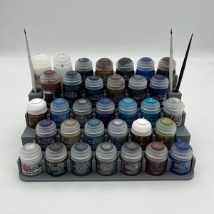 May include: A gray plastic paint rack with multiple rows of small paint bottles. The bottles are mostly blue and green, with some black and white bottles. The bottles are labeled "Citadel Colour".
