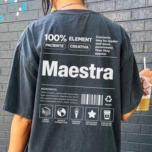 May include: A charcoal gray short-sleeve t-shirt featuring white graphic text on the back. The design includes the word Maestra, a barcode, and icons representing a book, globe, torch, star, and coffee cup with the text 100% ELEMENT PACIENTE CREATIVA.
