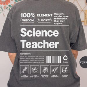 May include: Dark gray t-shirt with the words "Science Teacher" in large white letters. The shirt also features the text "100% Element" and science-related icons. The shirt's ingredients include love for hypotheses and endless curiosity.