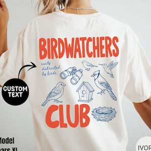 May include: Ivory-colored t-shirt with a red and blue graphic design. The design features the words "Birdwatchers Club" with illustrations of birds, binoculars, a birdhouse, and a nest. The text "easily distracted by birds" is also included.