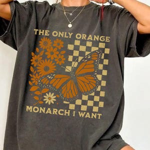 May include: Dark gray t-shirt featuring a monarch butterfly graphic with the text "THE ONLY ORANGE MONARCH I WANT". The design includes orange flowers and a checkered background, creating a vintage aesthetic.