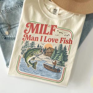 May include: Ivory-colored t-shirt with the text "MILF Man I Love Fish" in red and green. The graphic depicts a fishing scene with a large fish jumping out of the water. Light blue jeans and a tan hat are also in the image.