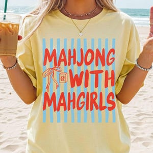 Peut inclure: Un t-shirt &agrave; manches courtes jaune p&acirc;le avec un imprim&eacute; graphique compos&eacute; de rayures verticales bleu clair et du texte rouge "MAHJONG WITH MAHGIRLS", accompagn&eacute; d'une petite illustration de ruban rouge et d'une tuile de mah-jong.