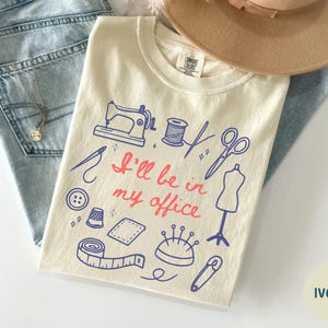 May include: Ivory-colored t-shirt with a sewing-themed design in blue and red, featuring a sewing machine, scissors, and the text "I'll be in my office." The shirt is paired with light-wash jeans and a tan hat.